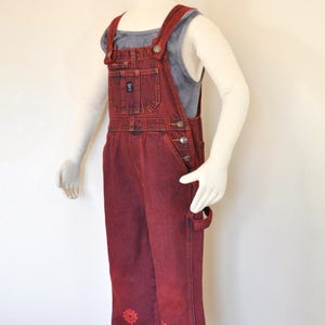 May include: Red denim overalls with a floral design on the lower legs. The overalls have a bib pocket, side pockets, and adjustable straps. A gray tank top is worn underneath. The overalls are displayed on a mannequin.