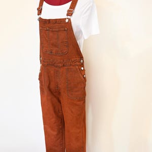 Puede incluir: Mono vaquero color óxido con tirantes ajustables y múltiples bolsillos. El mono se combina con una camiseta blanca de canalé. La prenda se exhibe en un maniquí sobre un fondo neutro, mostrando un estilo casual.