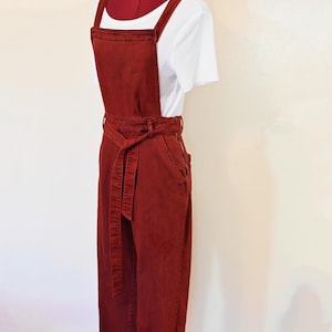 Puede incluir: Un mono vaquero de color óxido con cinturón a juego y perneras anchas. El mono tiene una parte superior tipo peto con tirantes ajustables y una camiseta blanca de canalé debajo. El conjunto se exhibe en un maniquí.