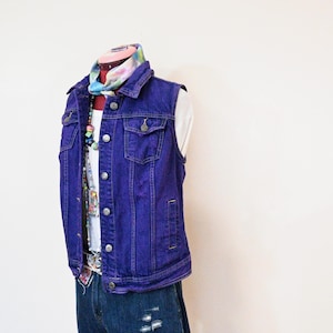 以下が含まれることがあります： マネキンに飾られた、シルバーのボタンが付いた紫色のデニムベスト。白いトップスの上に重ね着され、ダメージ加工のデニムショーツと組み合わされています。カラフルなスカーフとビーズのネックレスが、コーディネートにアクセントを加えています。