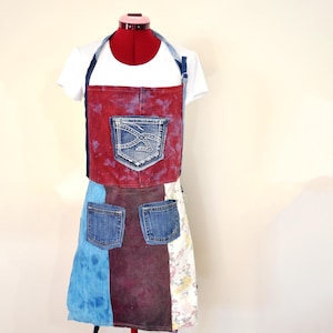 Op de afbeelding: Schort #46, Volwassen Small. Een patchwork schort gemaakt van gerecycled denim en andere stoffen. De schort heeft een rode denim top met een voorzak, twee onderste zakken en een veelkleurige rok. De schort wordt op een mannequin getoond.