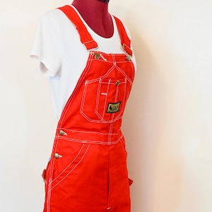 Könnte beinhalten: Leuchtend rote Latzhose mit weißen Nähten und messingfarbenen Beschlägen. Das Kleidungsstück wird auf einer Schaufensterpuppe mit einem weißen T-Shirt präsentiert. Ein kleines Patch ist auf der Latz-Tasche sichtbar.