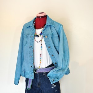 May include: An outfit comprising a light blue denim jacket, white t-shirt, and denim shorts. The ensemble includes a purple belt and colourful beaded necklaces. The shorts feature distressed details. The outfit is displayed on a mannequin.