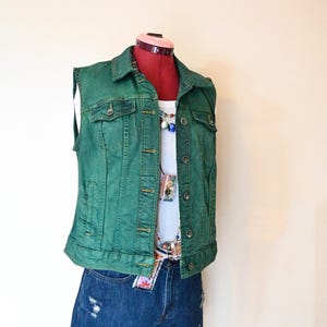 Op de afbeelding: Een groene spijkerweste met knopen, gedragen over een witte tanktop en een spijkerbroek. De outfit wordt getoond op een rode paspop. Een kleurrijke ketting en riem voegen accenten toe aan de ensemble. De shorts hebben distressed details.