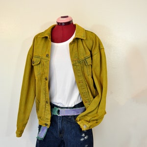 Op de afbeelding: Een mosterdgele denimjas met een wit T-shirt eronder en blauwe denim shorts met een paarse en groene riem.