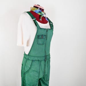 May include: A pair of green denim overalls featuring a front zipper closure, a chest pocket with a button flap, and two side pockets. The overalls are worn over a white short-sleeved ribbed shirt and a colorful tie-dye neck scarf.