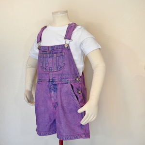 May include: A pair of purple denim overall shorts with a white t-shirt. The shorts have a classic bib pocket, side pockets, and adjustable straps. The outfit is displayed on a mannequin.