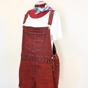 Könnte beinhalten: Rote Jeans-Latzshorts mit einem weißen T-Shirt darunter. Die Shorts haben eine Vordertasche und seitliche Knopfverschlüsse. Ein farbenfrohes Halstuch ist um den Hals der Schaufensterpuppe gebunden.