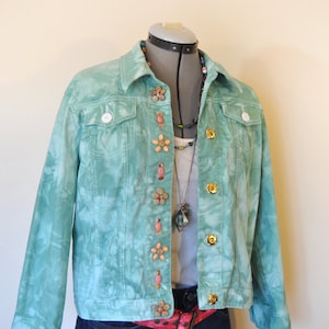 May include: A light teal denim jacket with a tie-dye effect, decorated with flower-shaped buttons and gold-coloured metal buttons. The jacket is displayed on a mannequin, paired with a white top, a pink belt, and dark blue jeans.