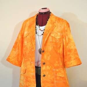 Puede incluir: Una blazer naranja teñida con nudos con mangas cortas, dos bolsillos delanteros y botones oscuros. La blazer se combina con una camiseta blanca, pantalones cortos de mezclilla y un collar de cuentas. El atuendo se exhibe en un maniquí.