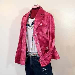 Puede incluir: Una chaqueta rosa teñida con tinte anudado con una camiseta blanca, pantalones cortos de mezclilla y collares de cuentas de colores. La chaqueta tiene botones y dos bolsillos delanteros. También se ve un cinturón estampado.