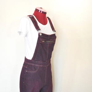 Puede incluir: Mono vaquero color ciruela con tirantes ajustables y un bolsillo delantero con cremallera. El mono se combina con una camiseta blanca, exhibido en un maniquí. El mono tiene un diseño clásico de cinco bolsillos.