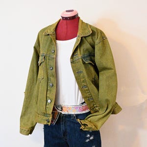 Op de afbeelding: Een groene spijkerjas met knopen, gedragen over een witte tanktop en een blauwe spijkerbroek. De outfit bevat een riem met patroon. De kleding wordt op een mannequin gepresenteerd.