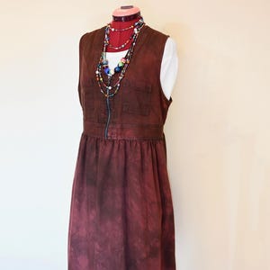 Peut inclure: Robe bordeaux sans manches avec fermeture éclair sur le devant et deux poches poitrine. La robe a une taille froncée et une jupe évasée. Un haut blanc est visible en dessous. Des colliers de perles colorées ornent l'encolure.