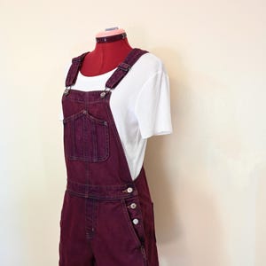 Könnte beinhalten: Burgunderfarbene Jeans-Latzhose mit Brusttasche, Seitentaschen und Knopfverschlüssen. Die Latzhose wird mit einem weißen Rippstrick-T-Shirt kombiniert. Das Outfit wird auf einer Schaufensterpuppe präsentiert.