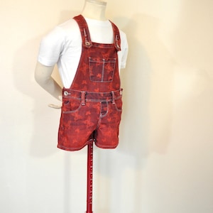 Puede incluir: Pantalones cortos con peto de mezclilla roja con estampado de mariposas, exhibidos en un maniquí. Los pantalones cortos tienen tirantes ajustables, bolsillos y cierre de botón. Se usa una camiseta blanca debajo. Es un artículo de moda de verano.