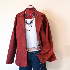 May include: A red denim jacket, white ribbed tank top, and denim shorts are displayed on a mannequin. The outfit is accessorized with a beaded necklace and a patterned belt. The shorts have distressed details.