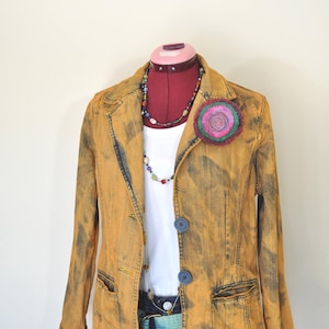 Puede incluir: Chaqueta vaquera amarilla mostaza con dos botones azules, una camisa blanca y un collar colorido. Un broche de flores decorativo está sujeto a la chaqueta. El atuendo se exhibe en un maniquí.