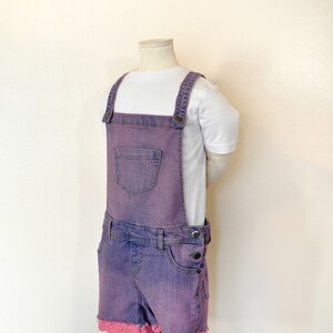 Puede incluir: Pantalón vaquero morado con tirantes y ribete de encaje rosa. El pantalón tiene un bolsillo delantero y tirantes ajustables.