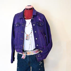 May include: A purple denim jacket with a white top, denim shorts, and a floral belt. The jacket has button closures and distressed details. A necklace with colorful beads and a pendant is also visible.