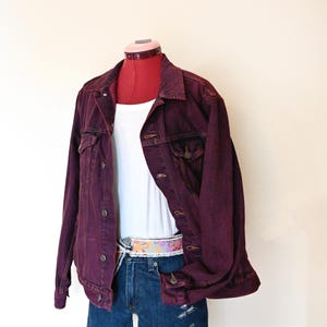 Puede incluir: Chaqueta vaquera burdeos, camiseta sin mangas blanca y shorts vaqueros. La chaqueta tiene botones y bolsillos. Se lleva un cinturón estampado de colores con los pantalones cortos. El conjunto se exhibe en un maniquí rojo.