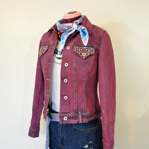 May include: A burgundy denim jacket with a light blue bandana and a beaded necklace. The jacket has rhinestones on the pockets and a distressed look. The jacket is paired with dark blue denim shorts.