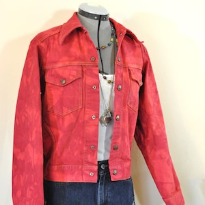 Puede incluir: Chaqueta vaquera roja con botones, dos bolsillos en el pecho y cuello. Combinada con una camiseta blanca, un collar y unos shorts vaqueros azules. Las medidas de la chaqueta no son visibles.