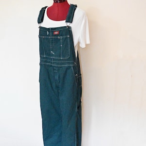Può includere: Salopette in denim blu scuro con spalline regolabili e multiple tasche, esposta su un manichino. La salopette è abbinata a una t-shirt bianca. Il logo Dickies è visibile sulla tasca della pettorina. L'outfit è esposto su un pavimento in legno.