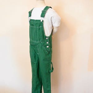 Può includere: Salopette in denim verde con cuciture bianche e bottoni argentati, abbinata a una maglietta bianca. La salopette ha spalline regolabili e più tasche. L'outfit è esposto su un manichino.