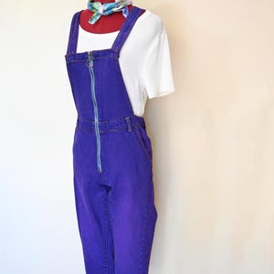 Könnte beinhalten: Eine lila Jeans-Latzhose mit hellblauem Reißverschluss und Trägern. Dazu ein weißes Kurzarm-Shirt und ein farbenfrohes Halstuch. Das Outfit wird auf einer Schaufensterpuppe präsentiert.