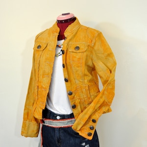 Puede incluir: Chaqueta amarillo mostaza con botones, usada sobre una camiseta blanca y pantalones cortos de mezclilla. La chaqueta tiene múltiples bolsillos y un aspecto desgastado. Un collar de cuentas de colores y un cinturón estampado completan el atuendo.