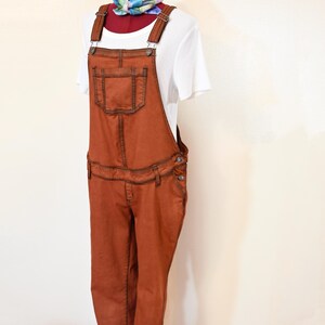 Puede incluir: Mono vaquero color óxido con costuras oscuras y un bolsillo delantero. El mono se combina con una camiseta blanca de manga corta y una bufanda colorida. El conjunto se exhibe en un maniquí.