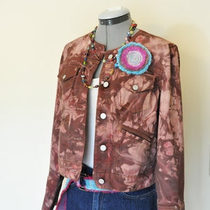 May include: A brown denim jacket with a tie-dye effect, paired with denim shorts. The jacket features silver buttons and pockets. A colorful beaded necklace and a large flower pin adorn the jacket. A blue and pink belt completes the look.