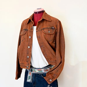 Puede incluir: Chaqueta vaquera color óxido con botones, camiseta blanca y shorts vaqueros desgastados. El conjunto se complementa con un cinturón estampado colorido. La chaqueta tiene un pequeño logo en el bolsillo.