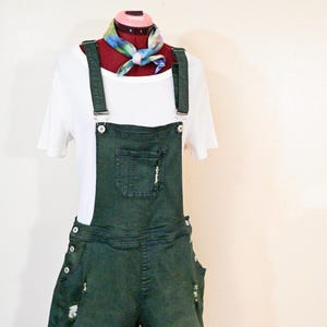 May include: Dark green denim overall shorts with distressed details, paired with a white t-shirt and a colorful scarf. The shorts have pockets and adjustable straps, displayed on a mannequin.