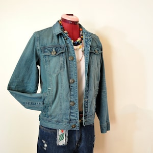 May include: A casual outfit comprising a denim jacket and shorts. The jacket is a mid-blue wash with button fastenings and pockets. The shorts are dark blue denim with distressed detailing. A white top and a colourful beaded necklace are also visible.