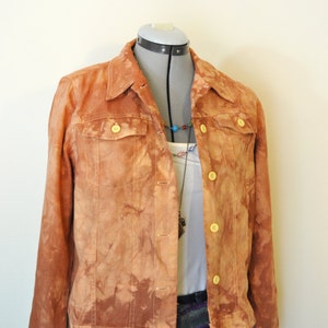 Puede incluir: Chaqueta vaquera naranja-marrón con efecto tie-dye, botones y dos bolsillos en el pecho. La chaqueta se muestra en un maniquí, combinada con una camiseta blanca y vaqueros azules. También se ve un collar de cuentas.