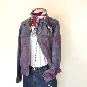 May include: A purple and gray tie-dye denim jacket with a white t-shirt, denim shorts, and a colorful scarf. The outfit is accessorized with a beaded necklace and a black belt. The jacket has two chest pockets and button closures.