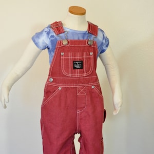 Op de afbeelding: Rode denim tuinbroek met witte stiksels en metalen hardware. De tuinbroek heeft een voorzak met een label met de tekst "Lakin Mickey". Een blauw tie-dye t-shirt wordt eronder gedragen.