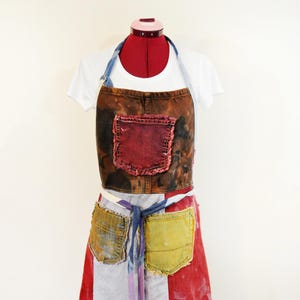 Op de afbeelding: Schort #53, Volwassen Medium, weergegeven op een mannequin. De schort heeft een patchwork-ontwerp met een bruine top, een rode zak en een rok met rode, witte en gele patches. Het heeft een blauwe denim nekband en taillebanden.