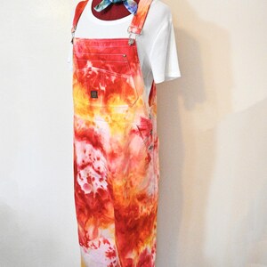 Puede incluir: Un mono teñido anudado en tonos rojos, naranjas y amarillos, exhibido en un maniquí. El mono tiene tirantes ajustables y bolsillos. Se usa una camiseta blanca debajo y una bufanda colorida atada alrededor del cuello del maniquí.