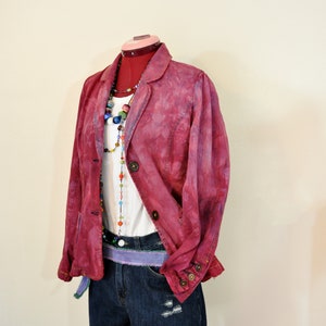 May include: A magenta-colored blazer with large brown buttons, layered over a white t-shirt and denim shorts. The outfit is accessorized with a beaded necklace and a purple belt. The shorts have distressed details.