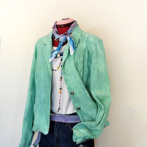 May include: A mint green blazer, white top, and denim shorts outfit. A patterned scarf is tied around the mannequin's neck, and a purple belt is worn at the waist. A colorful beaded necklace adds detail. The outfit is displayed on a red mannequin.