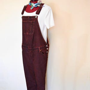 Könnte beinhalten: Burgunderfarbene Jeans-Latzhose mit verstellbaren Trägern und einer Vordertasche. Die Latzhose wird mit einem weißen T-Shirt und einem bunten Schal kombiniert. Die Latzhose hat seitliche Knopfverschlüsse und klassische Jeans-Taschendetails.