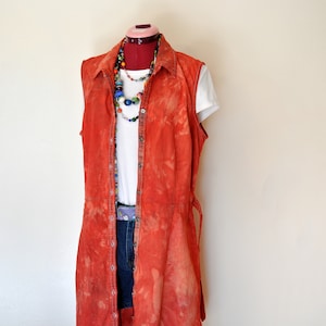 Könnte beinhalten: Ein orangefarbenes ärmelloses Hemdkleid mit Batik-Effekt, Knopfleiste vorne und einem weißen T-Shirt darunter. Das Outfit umfasst Jeansshorts, eine Perlenkette und einen lila Gürtel. Das Kleid wird auf einer Schaufensterpuppe präsentiert.