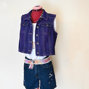 以下が含まれることがあります： 濃いインディゴ色のデニムベストに、デニムショートパンツと白いトップスを合わせたコーディネート。フローラルベルトと重ね着のネックレスがアクセントとなり、カジュアルでスタイリッシュなアンサンブルを演出。ショートパンツにはダメージ加工が施されています。