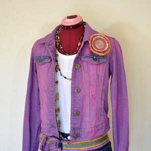 May include: A purple denim jacket with a colorful embroidered patch on the left shoulder. The jacket has a white t-shirt underneath and a brown and gold belt.