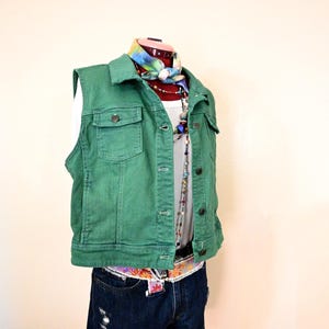 Op de afbeelding: Een groene spijkerstof vest met knoopsluitingen en twee borstzakken. Het vest wordt gecombineerd met een witte top, een spijkerbroek en een kleurrijke riem. Een sjaal met patroon en kralenkettingen maken de outfit compleet, gepresenteerd op een mannequin.