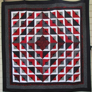 以下が含まれることがあります： 赤、黒、白、グレーの幾何学模様の手作りキルト。キルトは、正方形と三角形に囲まれた中央のダイヤモンドデザインが特徴です。黒い縁があり、壁に掛けられています。