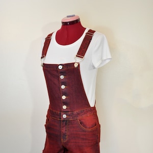 Könnte beinhalten: Rote Jeans-Latzhose-Shorts mit weißen Knöpfen und ausgefransten Kanten. Die Shorts werden mit einem weißen T-Shirt kombiniert. Die Latzhosenträger haben ein Muster. Das Outfit wird auf einer Schaufensterpuppe präsentiert.